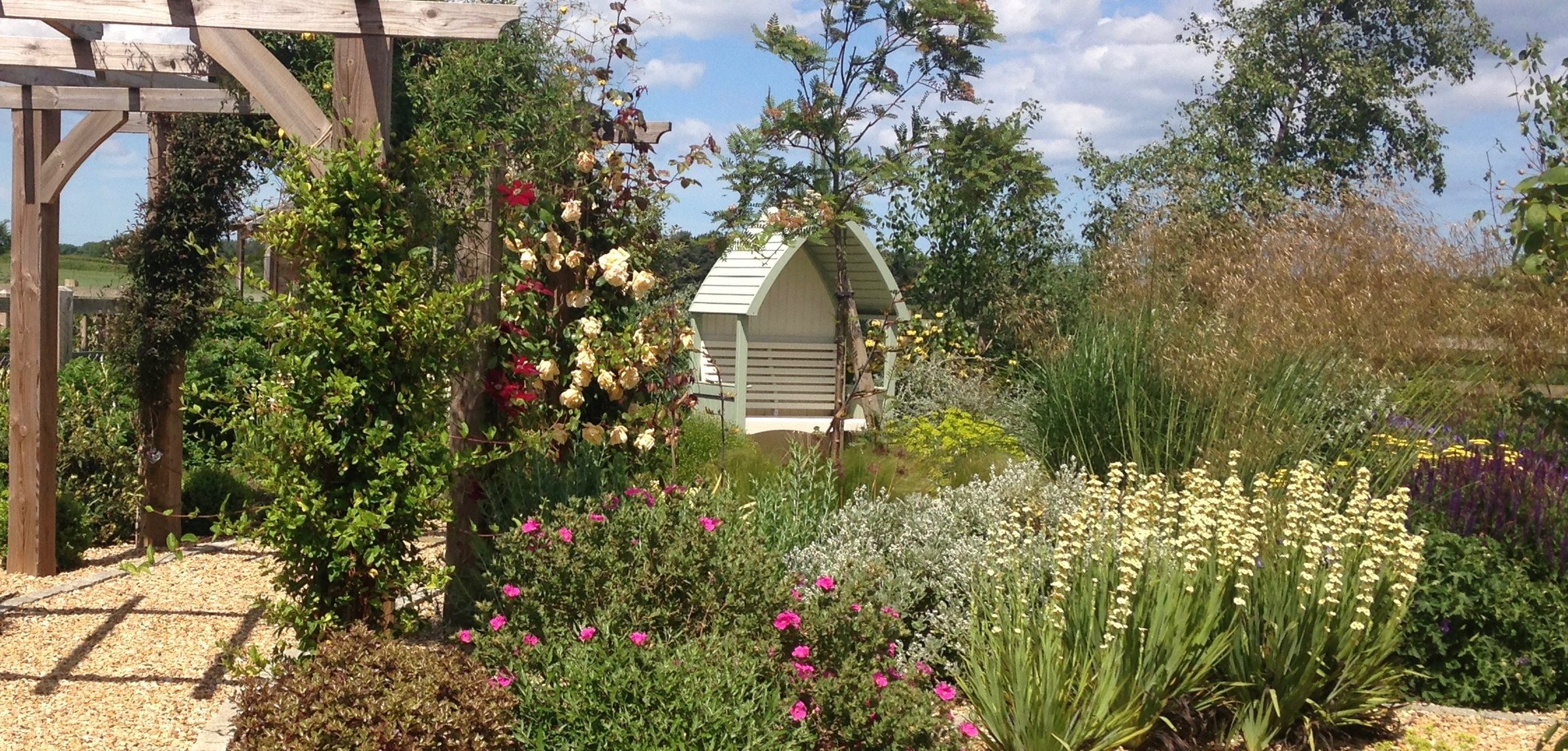 An oak pergola over a gravel path, framed by salvias and lavender under a summer sky